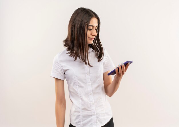 Молодая девушка в белой рубашке смотрит на экран своего мобильного телефона с улыбкой на лице, стоящей над белой стеной
