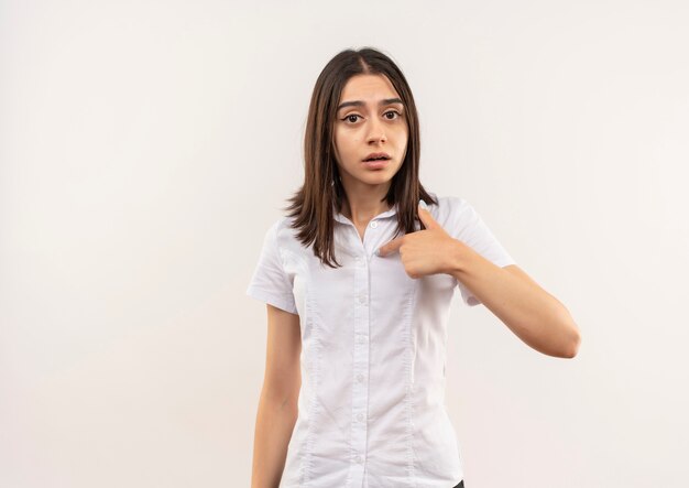 Young girl in white shirt pointing to herself looking confused standing over white wall