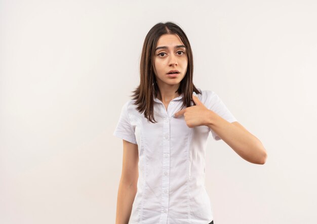 Молодая девушка в белой рубашке, указывая на себя, выглядит смущенной, стоя над белой стеной