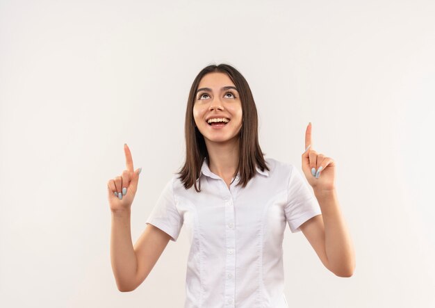 Молодая девушка в белой рубашке, указывая вверх указательными пальцами, счастливая и взволнованная, стоя у белой стены