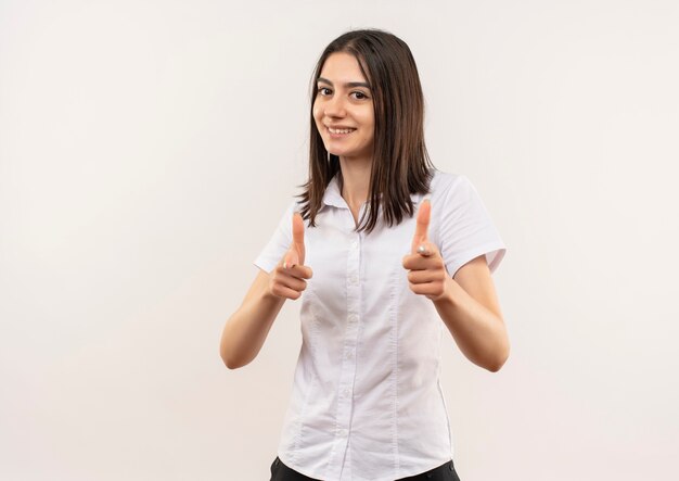 Молодая девушка в белой рубашке указывая указательными пальцами вперед, весело улыбаясь, стоя над белой стеной