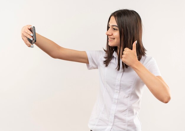 Young girl in white shirt taking selfie using her mobile smiling confident showing thumbs up to the front standing over white wall