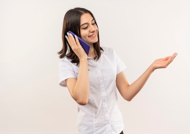 Молодая девушка в белой рубашке разговаривает по мобильному телефону с улыбкой на лице, стоящей над белой стеной