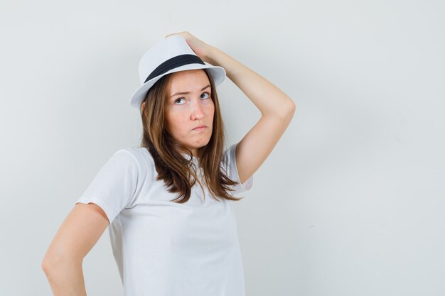 Young girl in white t-shirt, hat holding hand on head and looking pensive , front view.