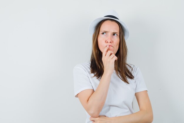 Young girl in white t-shirt, hat holding hand on lips and looking hesitant , front view.