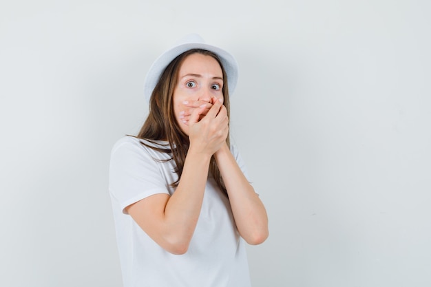 Young girl in white t-shirt, hat holding hands on mouth and looking scared , front view.