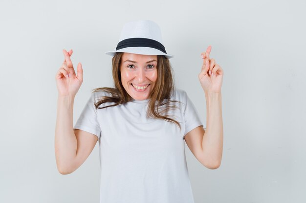 Young girl in white t-shirt, hat keeping fingers crossed and looking cheerful , front view.