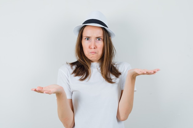 Young girl in white t-shirt, hat making scales gesture and looking confused , front view.