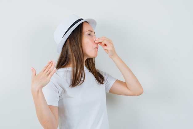 Young girl in white t-shirt, hat pinching nose due to bad smell and looking disgusted , front view.