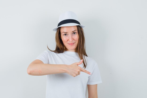 Young girl in white t-shirt, hat pointing down and looking confident , front view.