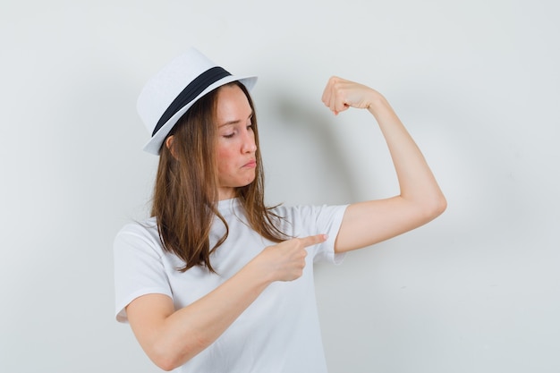 Young girl in white t-shirt, hat pointing at muscles of arm and looking proud , front view.