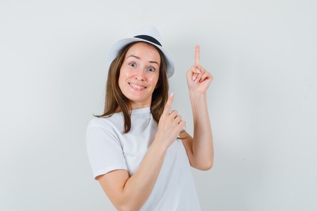 Young girl in white t-shirt, hat pointing up and looking blissful , front view.