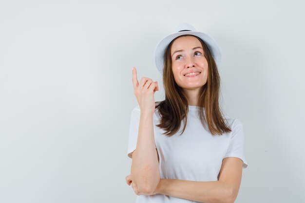 Young girl in white t-shirt, hat pointing up and looking hopeful , front view.