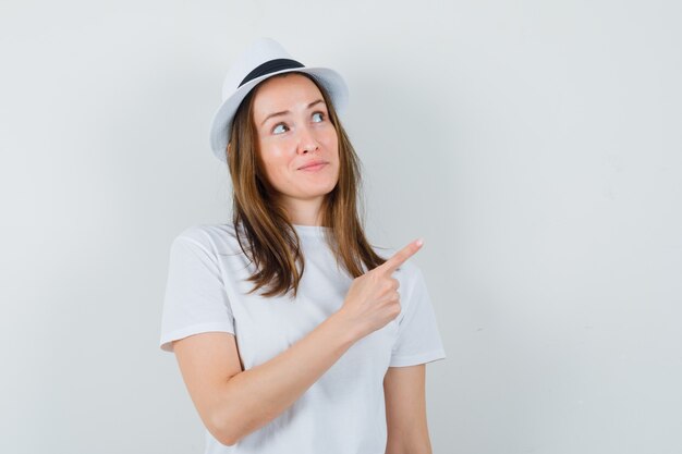 Young girl in white t-shirt, hat pointing at upper right corner and looking focused , front view.