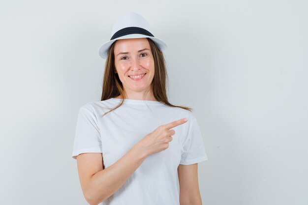 Young girl in white t-shirt, hat pointing at upper right corner and looking glad , front view.