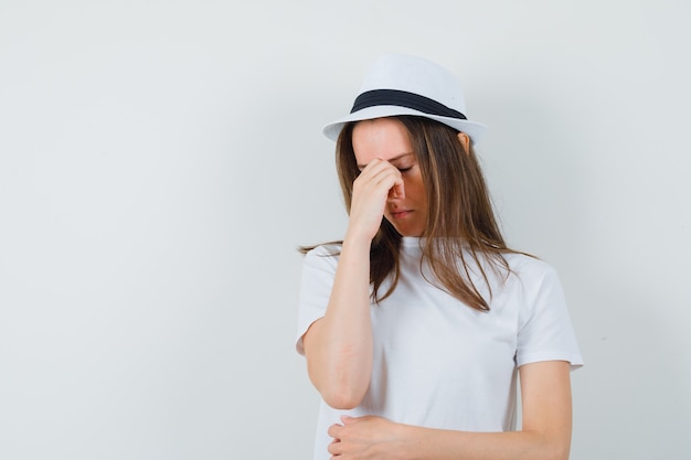 Young girl in white t-shirt, hat rubbing eyes and nose and looking upset , front view.