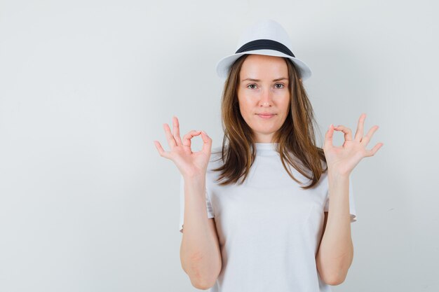 Young girl in white t-shirt, hat showing ok gesture and looking confident , front view.