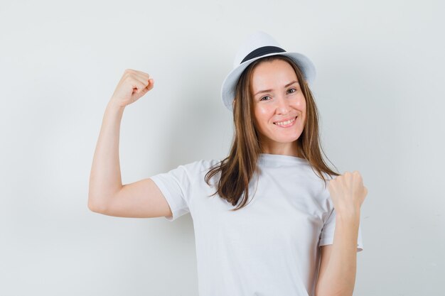 Young girl in white t-shirt, hat showing winner gesture and looking merry , front view.