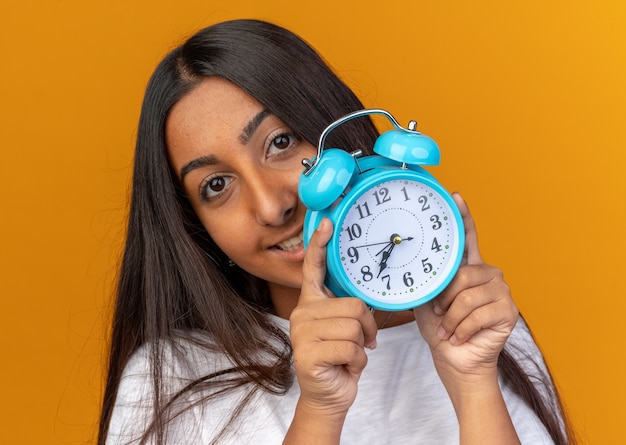Young girl in white t-shirt holding alarm clock looking at camera with happy face and smiling 