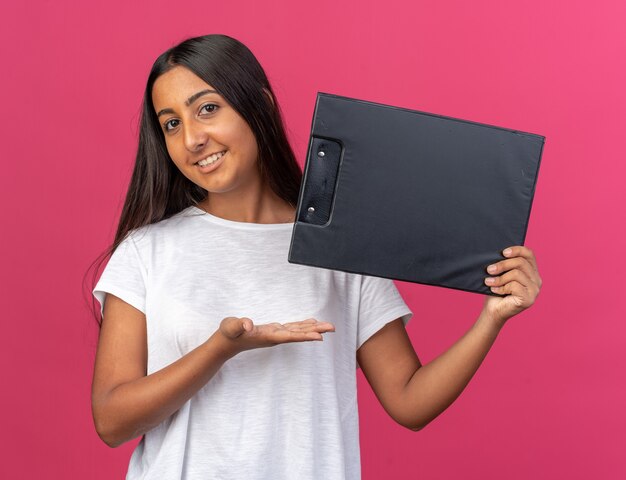 Young girl in white t-shirt holding clipboard looking at camera smiling cheerfully presenting with arm of hand standing over pink background