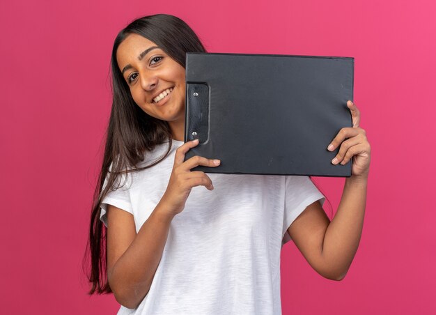 Young girl in white t-shirt holding clipboard looking at camera smiling cheerfully 