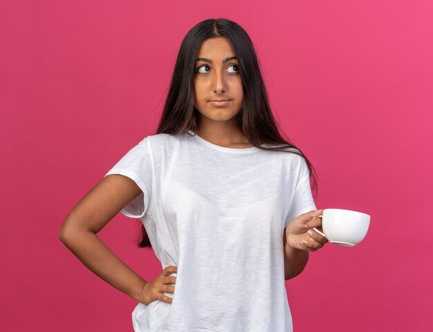 Молодая девушка в белой футболке держит чашку кофе с озадаченным взглядом, стоя над розовым