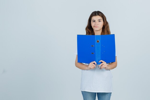 Young girl in white t-shirt holding folder, curving lips and looking disappointed , front view.