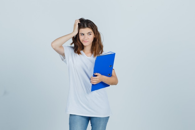 Young girl in white t-shirt holding folder, keeping hand on head and looking frustrated , front view.