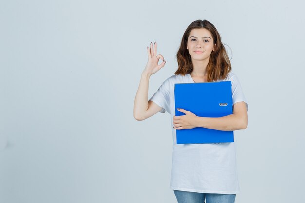 Young girl in white t-shirt holding folder, showing ok gesture and looking jolly , front view.