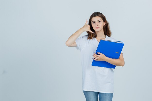 Молодая девушка в белой футболке держит папку, показывает жест телефона и выглядит полезным, вид спереди.