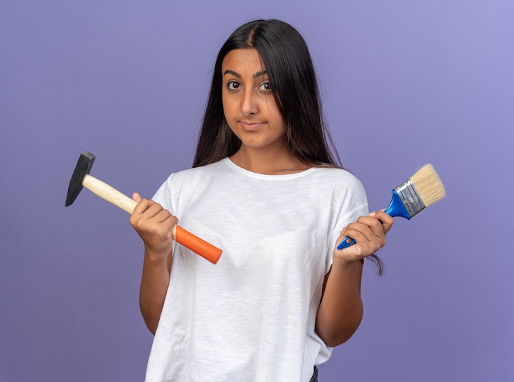 मुफ़्त फोटो young girl in white t-shirt holding hammer and paint brush looking at camera with confident smile on face standing over blue