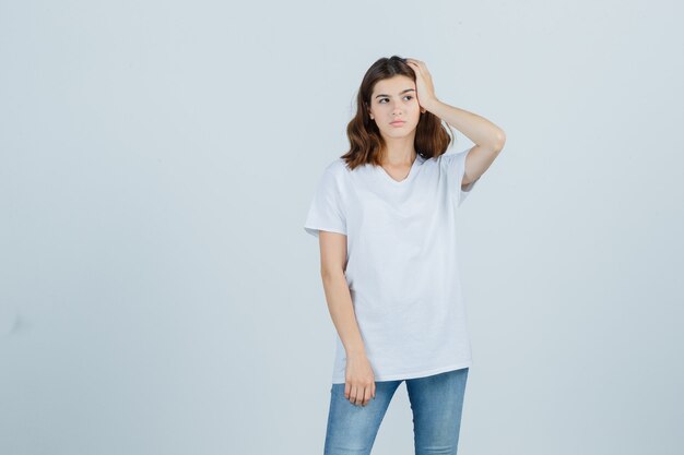 Young girl in white t-shirt holding hand on head and looking thoughtful , front view.