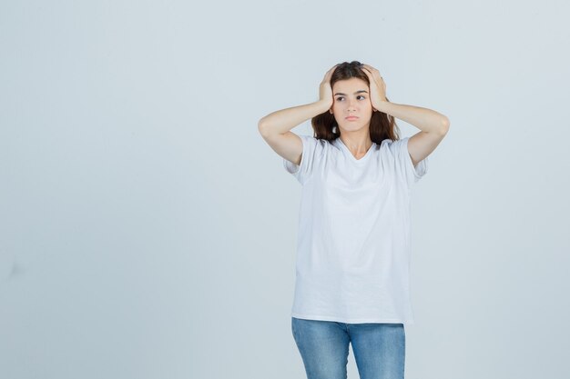 Young girl in white t-shirt holding hands on head and looking thoughtful , front view.