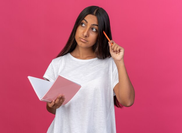 ピンクの背景の上に立っている鉛筆思考で困惑した引っかき頭を脇に見ているノートと鉛筆を保持している白いTシャツの少女