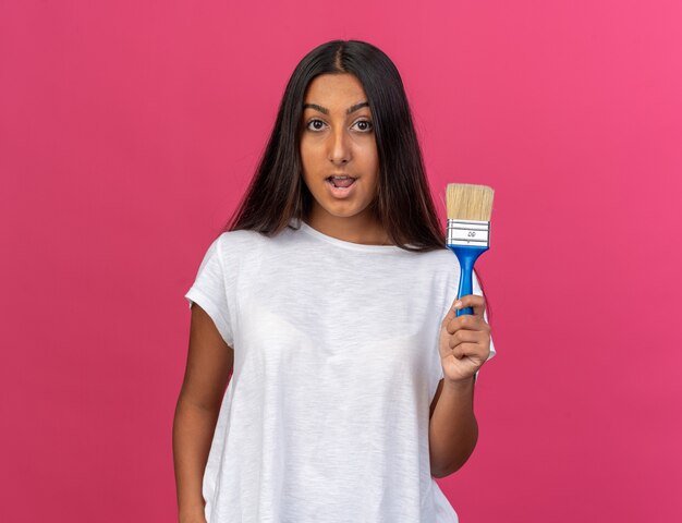 Young girl in white t-shirt holding paint brush looking at camera happy and surprised standing over pink