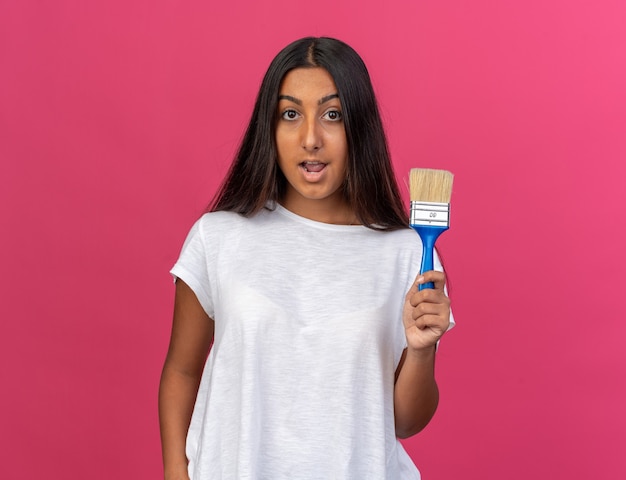 Young girl in white t-shirt holding paint brush looking at camera happy and surprised standing over pink