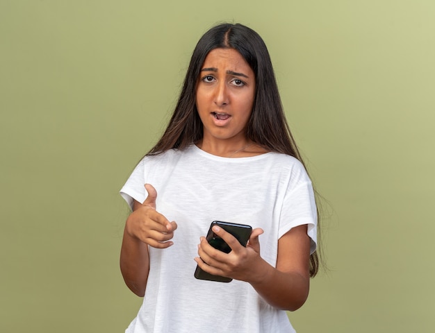 Young girl in white t-shirt holding smartphone looking at camera being confused and very anxious 