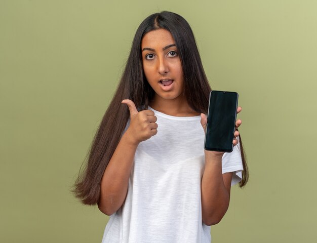 Young girl in white t-shirt holding smartphone looking at camera smiling confident showing thumbs up 