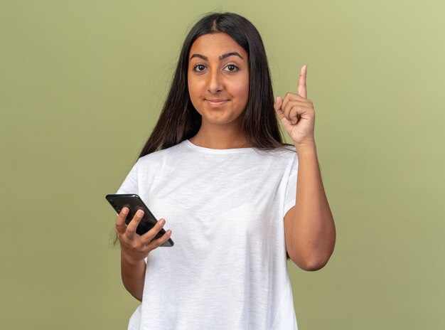 Молодая девушка в белой футболке держит смартфон, глядя в камеру с улыбкой на умном лице