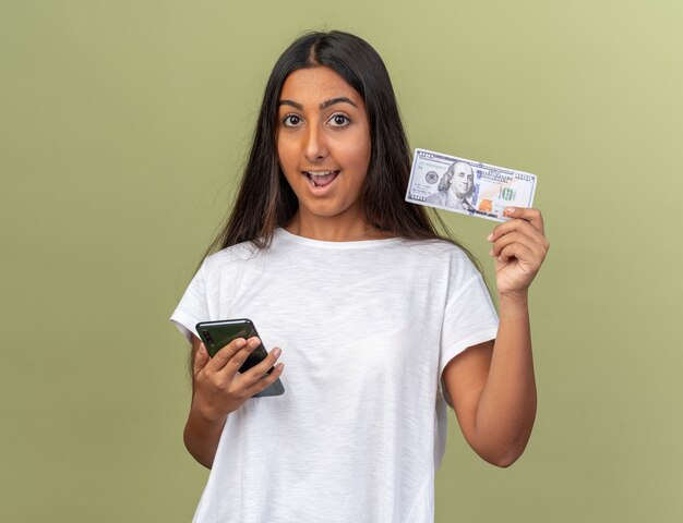 Young girl in white t-shirt holding smartphone showing cash looking at camera happy and surprised 