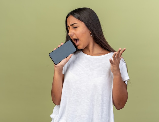 Young girl in white t-shirt holding smartphone using like a microphone singinh having fun standing over green background