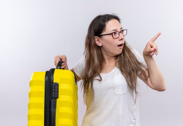 Young girl in white t-shirt holding travel suitcase  looking aside surprised and amazed pointing with index finger to something 