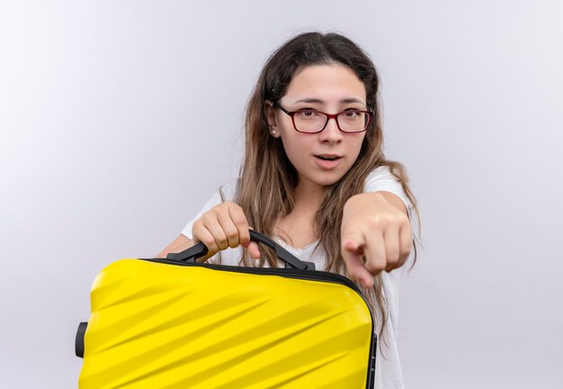 カメラに人差し指で指して驚いて見える旅行スーツケースを保持している白いTシャツの少女