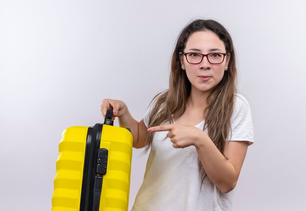 Young girl in white t-shirt holding travel suitcase  pointing with finger to it displeased 