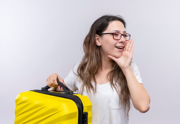 Giovane ragazza in maglietta bianca che tiene la valigia di viaggio gridando o chiamando qualcuno con la mano vicino alla bocca