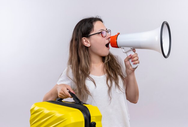 Молодая девушка в белой футболке держит дорожный чемодан и кричит в мегафон
