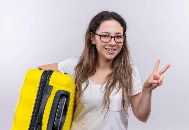 Young girl in white t-shirt holding travel suitcase  smiling cheerfully showing victory sign or two number 