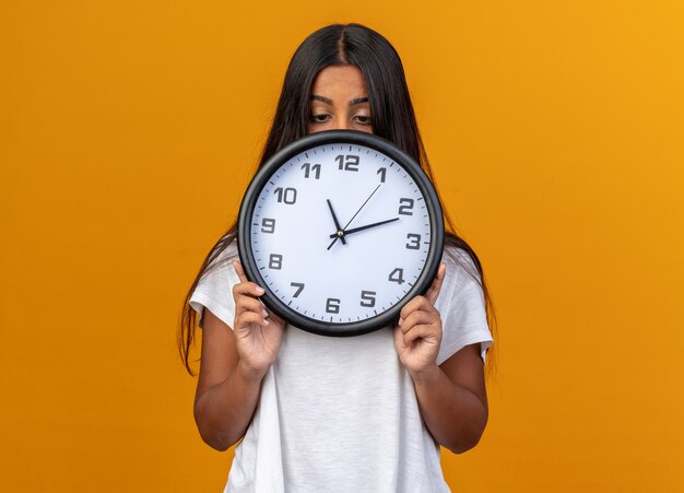 Young girl in white t-shirt holding wall clock hiding her face behind it standing over orange background