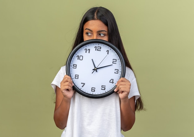 Young girl in white t-shirt holding wall clock looking aside with serious face standing over green background