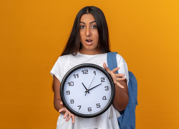 Young girl in white t-shirt holding wall clock looking at camera amazed and surprised standing over orange background